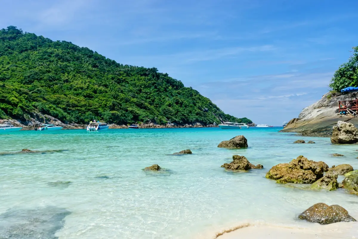 Rocky shoreline with turquoise water and boats anchored in the bay, surrounded by a lush green hillside at Banana Beach, one of the most scenic Phuket beaches.