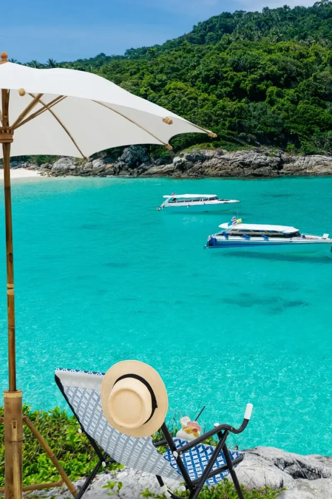 A sun hat resting on a beach chair beneath an umbrella, overlooking turquoise water and speedboats at Banana Beach, a popular spot among Phuket beaches.