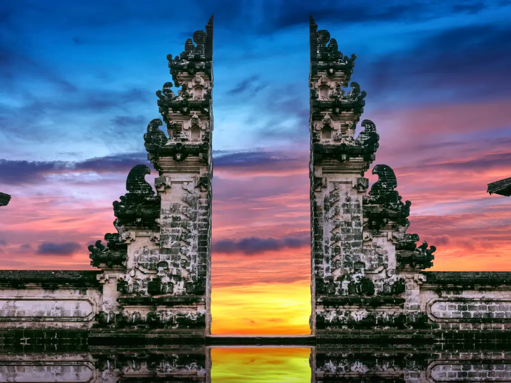 Lempuyang Temple sunset in Bali, Indonesia, showcasing its intricately carved gate against a vibrant sunset sky with hues of blue, purple, pink, and yellow. The gate's reflection in the water below adds to the serene and picturesque scene.