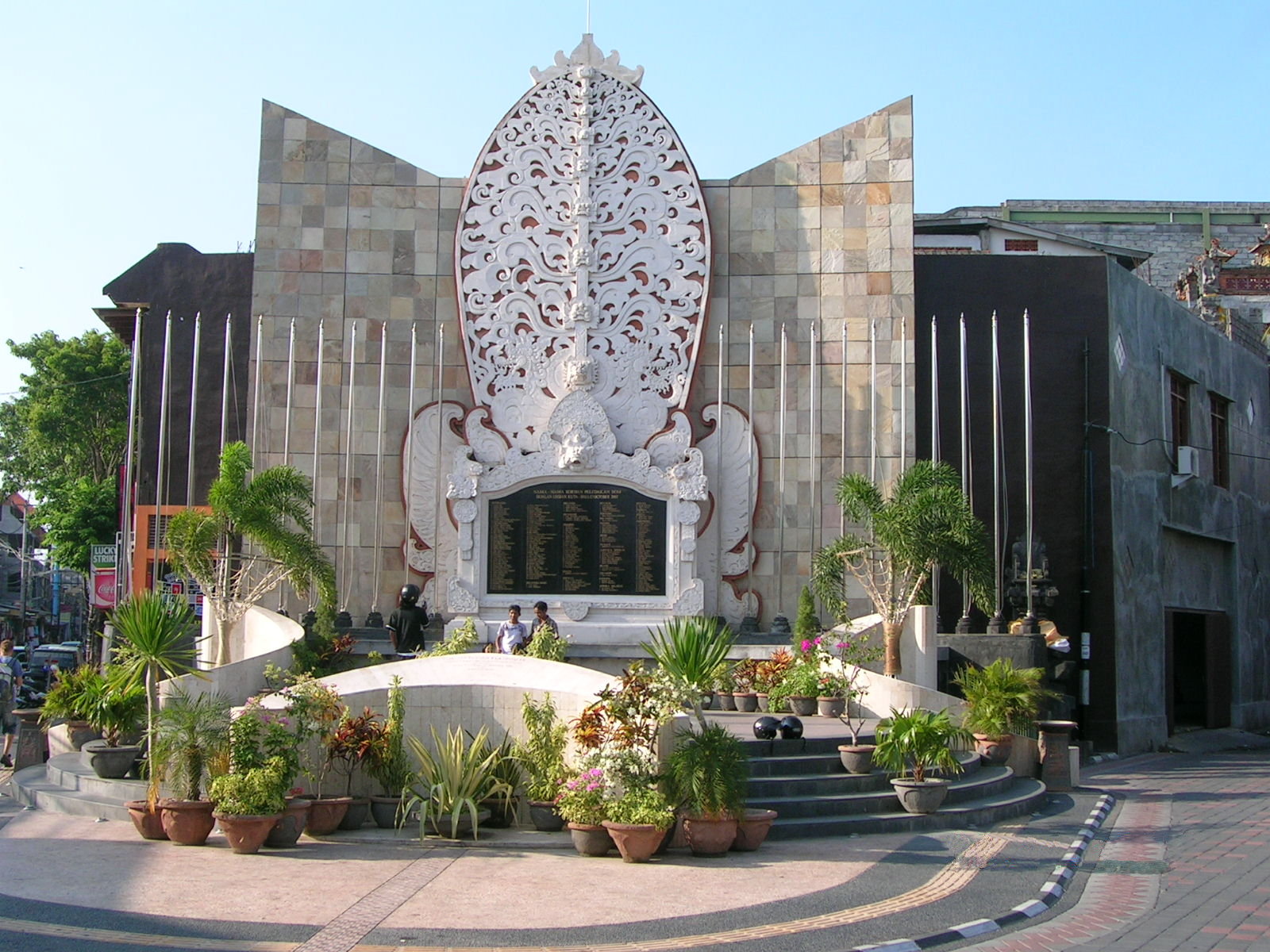 This image depicts the Bali Bombing Memorial in Kuta, Bali, Indonesia. The centerpiece features a carved stone monument listing the names of the victims surrounded by a beautifully landscaped area with potted plants and flags.