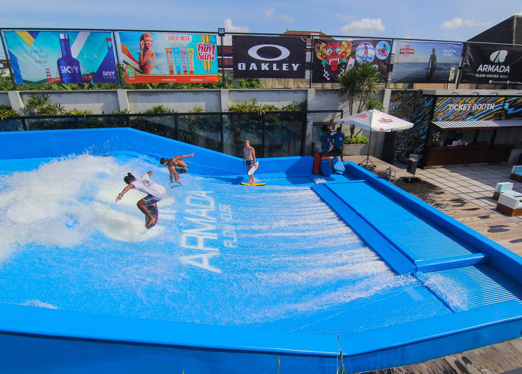 A vibrant artificial surfing wave pool at Armada Flow House Bali. Two people are riding the waves on bodyboards, while another stands nearby preparing to surf.