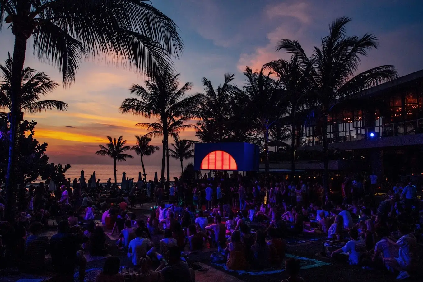 A lively evening scene at Potato Head Beach Club in Bali, with a crowd gathered under swaying palm trees during a vibrant sunset.