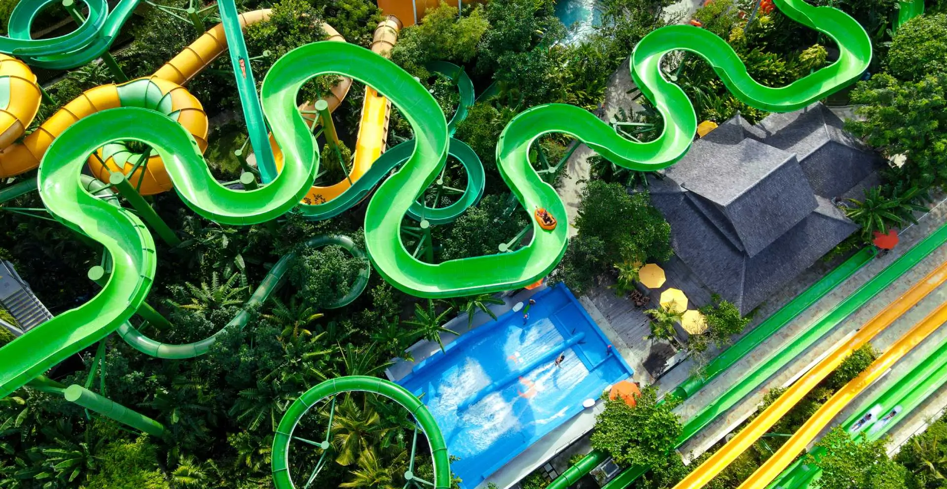 A vibrant aerial view of Waterbom Park in Bali, featuring winding green water slides that snake through lush tropical greenery. 