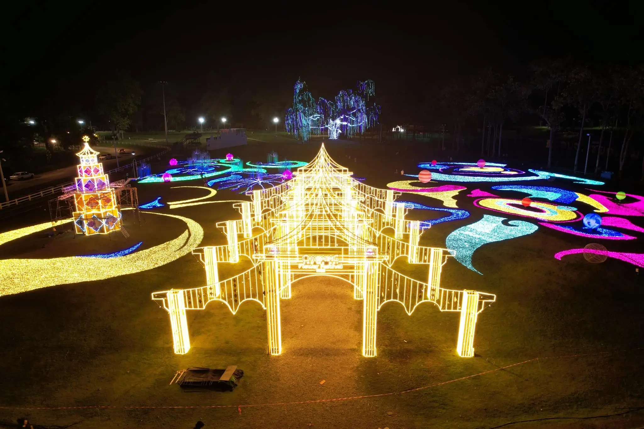 A vibrant aerial view of an illuminated park at night, showcasing glowing golden arches, a multi-tiered tower, and swirling patterns of colorful LED lights in blue, pink, and yellow.