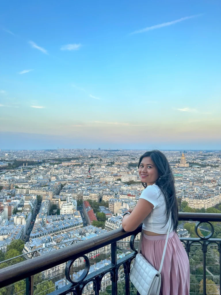 A woman smiling back at the camera, overlooking the panoramic view of Paris from the second floor of the Eiffel Tower, with the evening lights beginning to twinkle in the city below.
