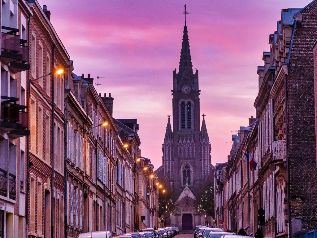 Dusk falls on a quiet street in Amiens, France, with the silhouette of the gothic Sainte-Anne church spire against a purple sky, and street lights casting a warm glow. This peaceful town atmosphere exemplifies the serene experiences of an easy day trip from Paris.