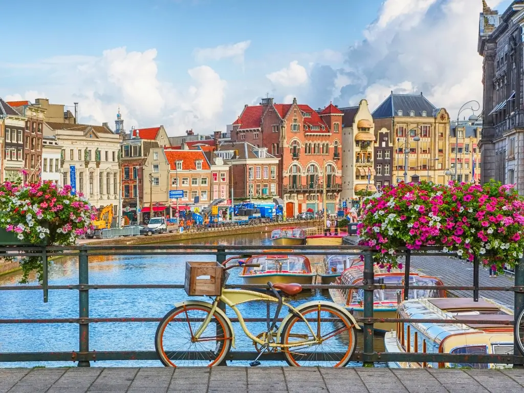 Colorful waterfront scene in Amsterdam with vibrant flowers in the foreground, a bicycle leaning against a fence, and historic Dutch buildings. Amsterdam is a favorite for those seeking the best day trips from Paris by train to other countries.