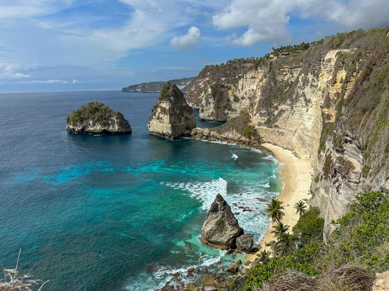A scenic view of Diamond Beach in Bali, featuring turquoise waters, dramatic limestone cliffs, and several jagged rock formations rising from the sea. The white sandy beach is lined with palm trees and bordered by steep, rugged cliffs under a partly cloudy sky.