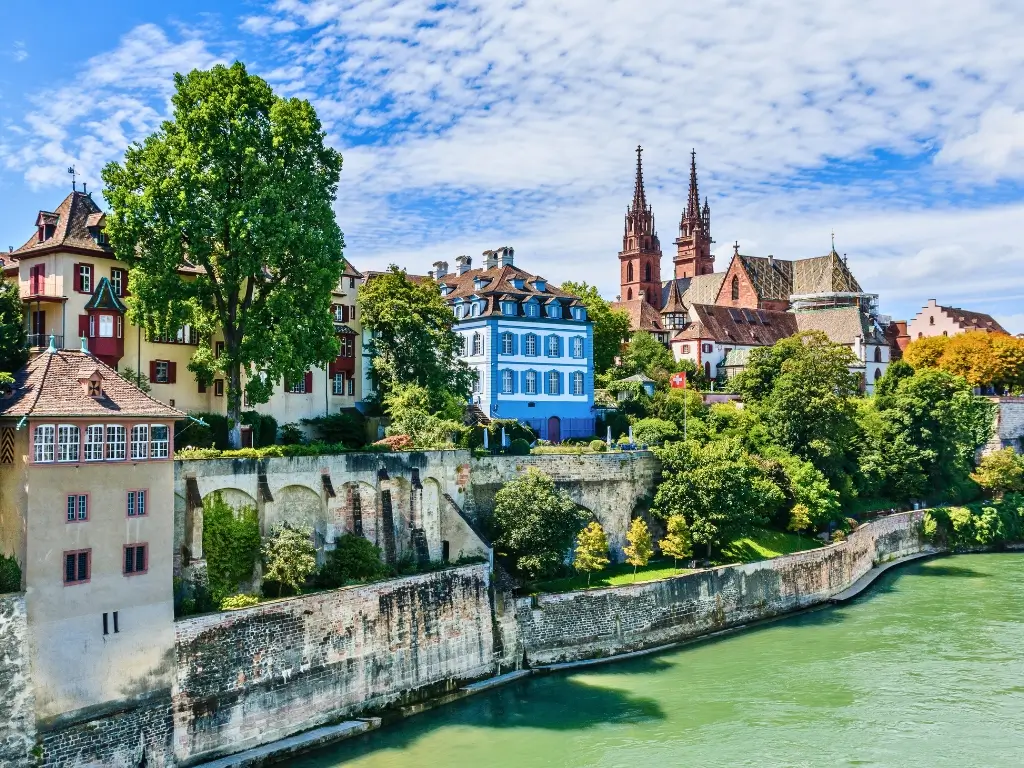 Historic and colorful riverside buildings in Basel, Switzerland, with the Basel Minster cathedral's spires standing out against the blue sky. This peaceful riverside view is representative of the sights encountered on the best day trips from Paris by train to other countries.
