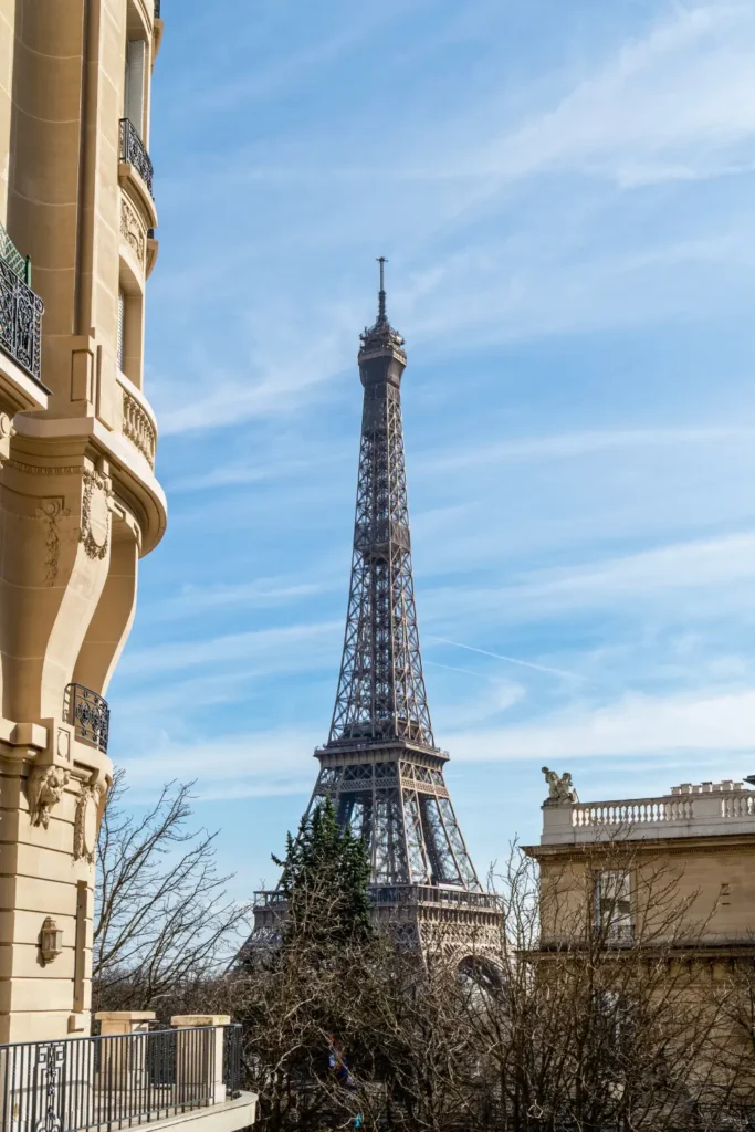 View of Eiffel Tower from Avenue de Camoëns, framed by classical Parisian architecture with ornate balconies, set against a clear blue sky.