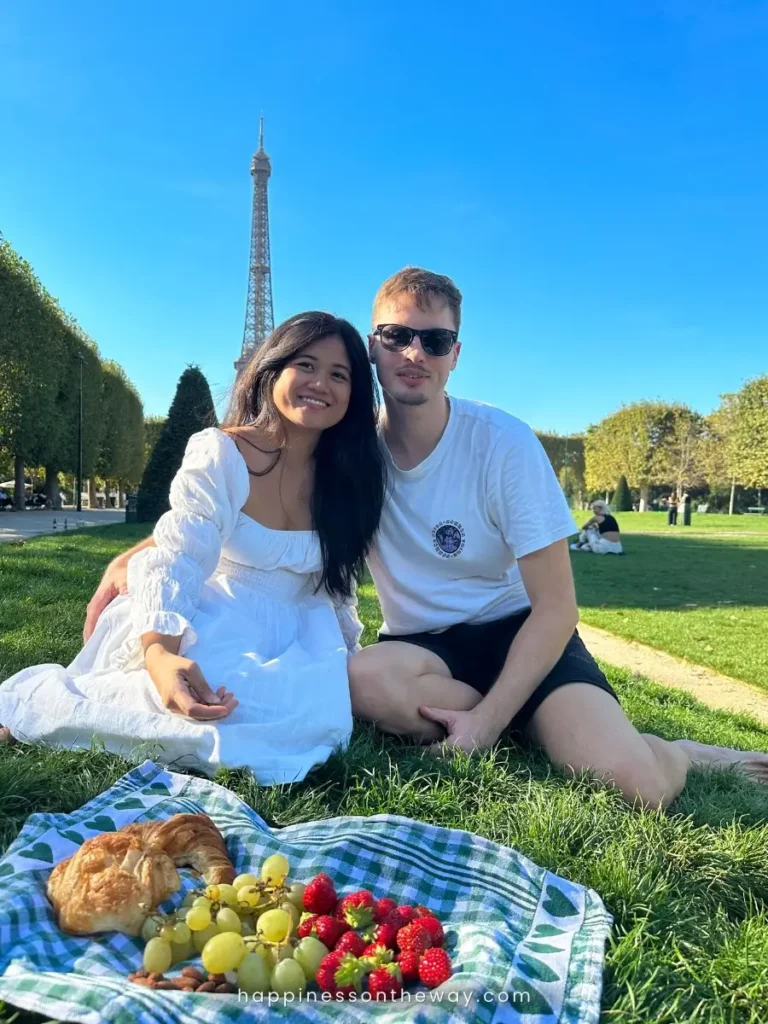 Us enjoying a picnic on the Champ de Mars with the best views of Eiffel Tower in the background. We're hapily seated on the grass with a spread of fruits, pastries, and nuts on a checkered cloth.