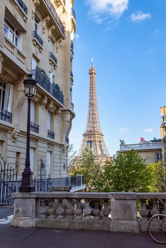 The majestic Eiffel Tower viewed from Avenue de Camoëns, lined with traditional Parisian lampposts and wrought-iron fences amidst green trees, on a sunny day.