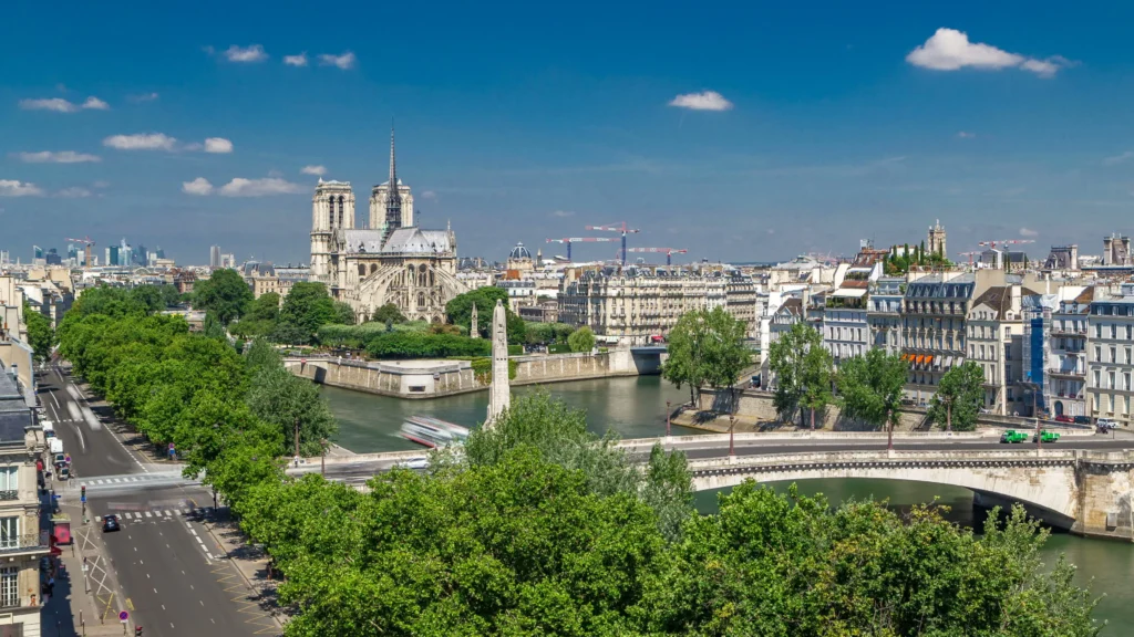 Panoramic view of Paris from the rooftop of Institut du Monde Arabe, featuring Notre-Dame Cathedral and the Seine River, with lush tree-lined streets and traditional Parisian buildings under a blue sky with scattered clouds.