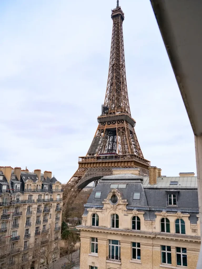 View of the Eiffel Tower rising above Parisian apartment buildings, as seen from a hotel balcony, with overcast skies above.