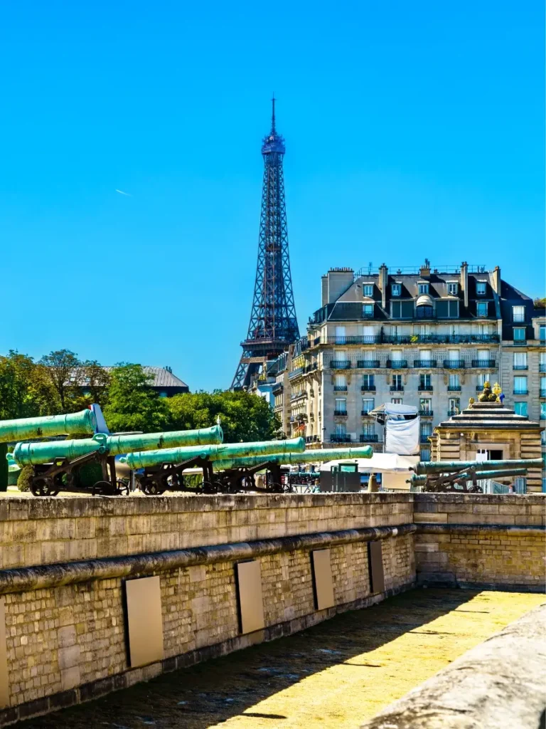 View from Les Invalides in Paris, featuring a row of antique cannons on display, with the Eiffel Tower rising behind classic French Haussmann-style buildings under a clear blue sky.