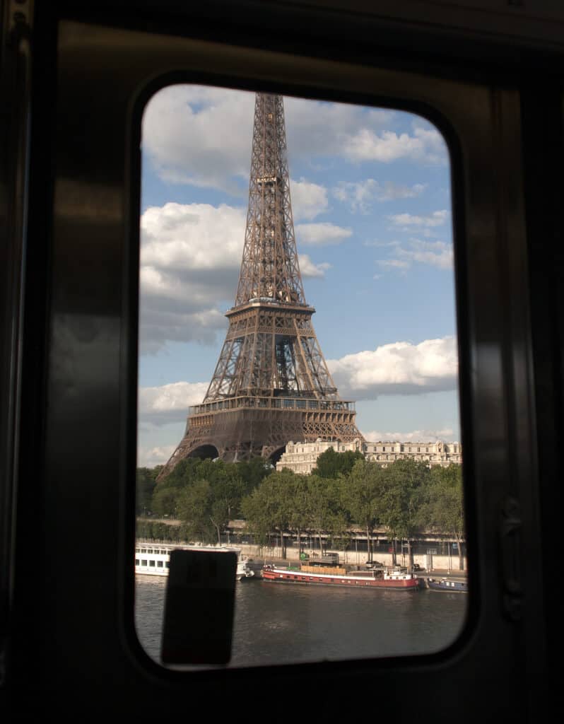View of the Eiffel Tower framed by the window of a metro train on line 6, with the Seine River and a passing barge in the foreground, on a day with scattered clouds in the sky.