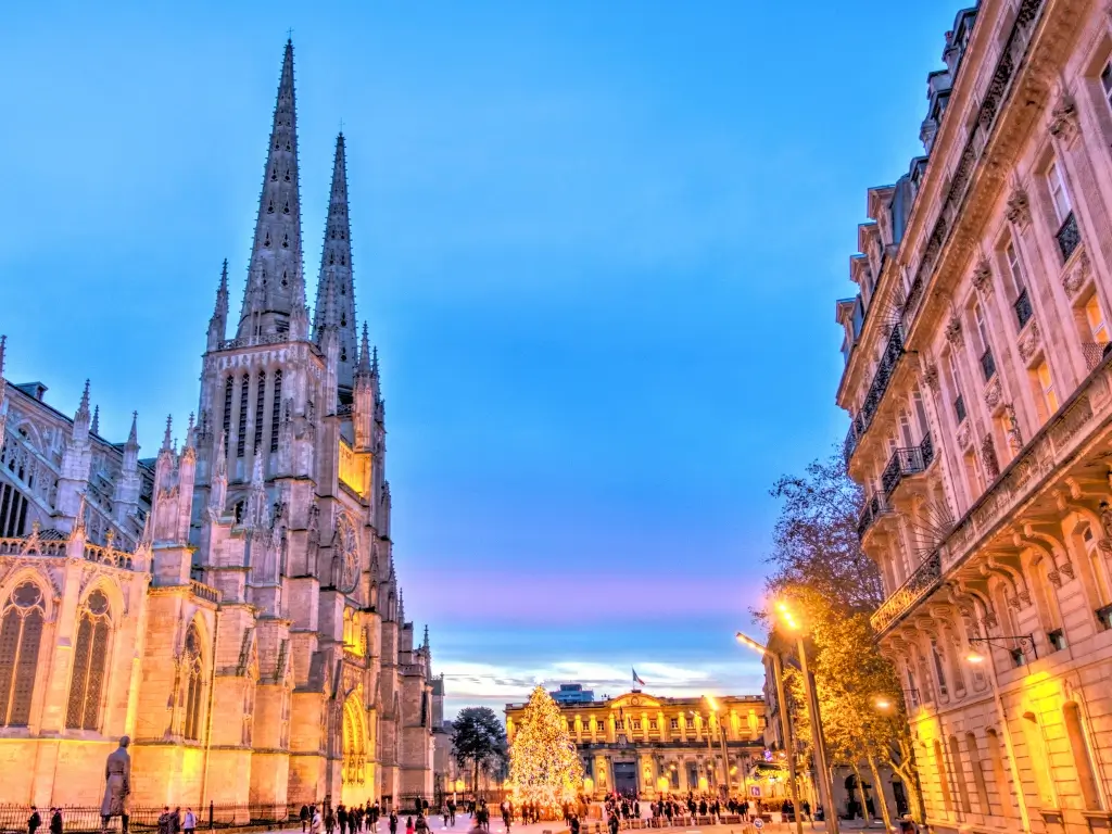 The gothic spires of Bordeaux Cathedral stand out against a twilight sky, with the warm glow of street lights illuminating the bustling square below. A captivating scene for those visiting Bordeaux, a gem among the best day trips from Paris by train.