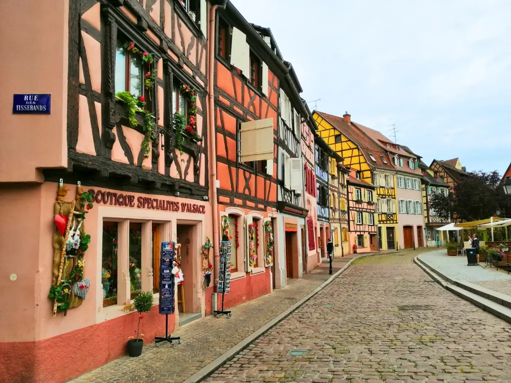 A quaint cobblestone street in Colmar, lined with traditional half-timbered houses painted in vibrant colors and adorned with flowers. Colmar, with its picturesque Alsace architecture, is a delightful day trip from Paris by train.
