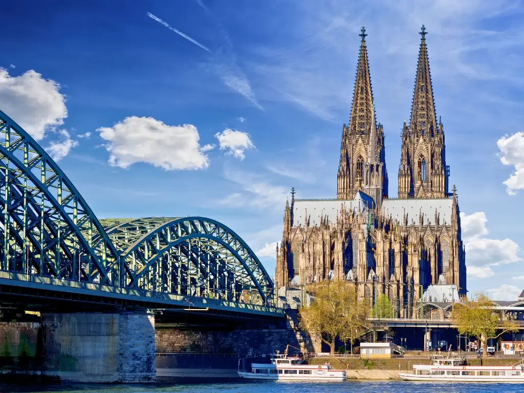 The Cologne Cathedral stands tall beside the Hohenzollern Bridge over the Rhine River in Cologne, Germany, with a tourist boat cruising by. This iconic Gothic church with its dual spires is a highlight of the best day trips from Paris by train to other countries.