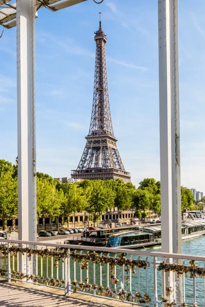 Eiffel Tower viewed through the structural beams of Debilly Footbridge with lock-covered railings and the Seine River below, showcasing a blend of Parisian engineering and romance.