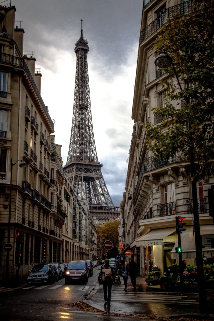 Dramatic view of the Eiffel Tower from Rue de Monttessuy on a cloudy day, with the tower's intricate metalwork standing out against the overcast sky, and the wet street reflecting the bustling Parisian life with pedestrians and cars.