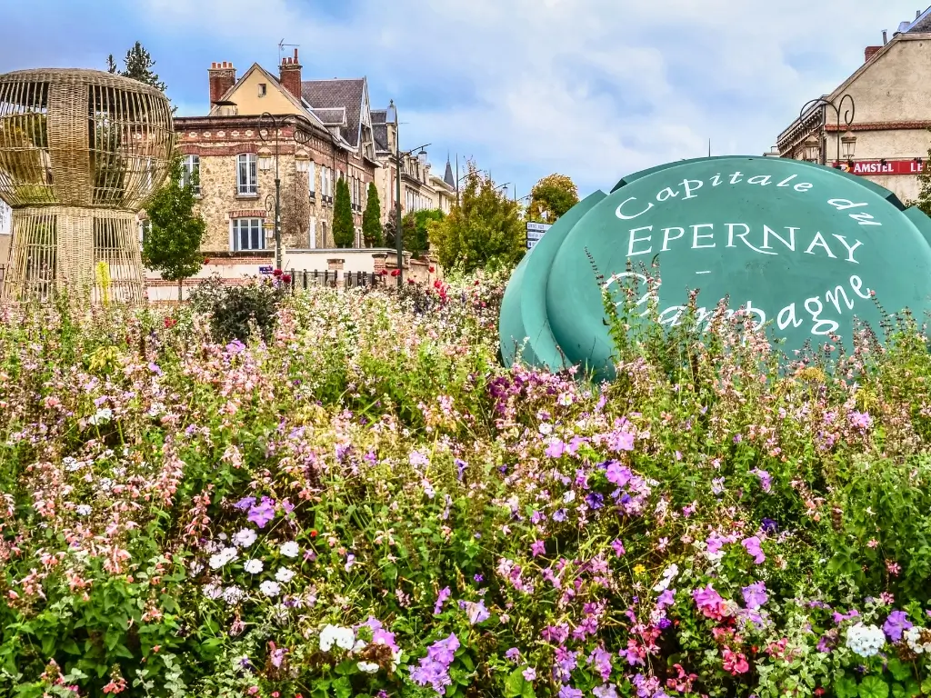 A colorful display of wildflowers in the foreground with a large, green sign reading 'Capitale de EPERNAY - Champagne' in the background, indicating Epernay's status as the heart of the Champagne region. This town is a celebrated day trip from Paris by train for those interested in champagne tasting and vineyard tours.