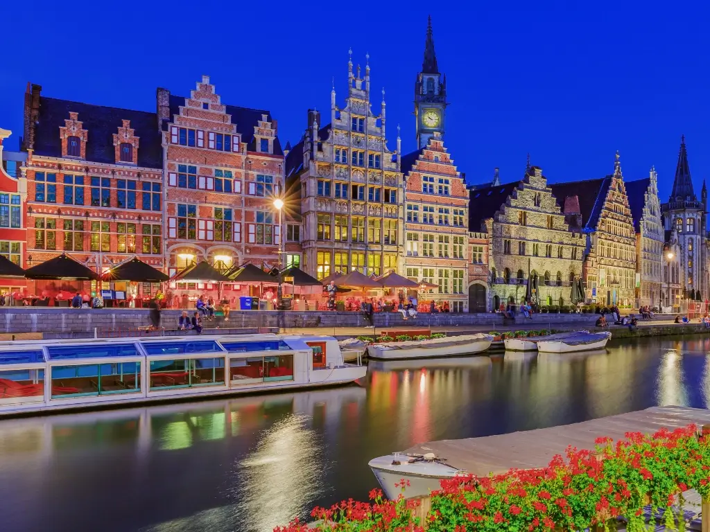 Twilight over the Graslei, the historic city center of Ghent, Belgium, with medieval buildings illuminated against the night sky and reflections on the calm river. This vibrant scene highlights the cultural experiences accessible on the best day trips from Paris by train.