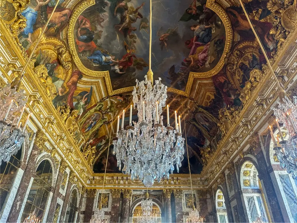 Inside the opulent Hall of Mirrors at the Palace of Versailles, with its grand crystal chandeliers, ornate gilded sculptures, and ceiling frescoes depicting classical scenes. Versailles is a splendid example of French grandeur and a famous day trip from Paris by train.