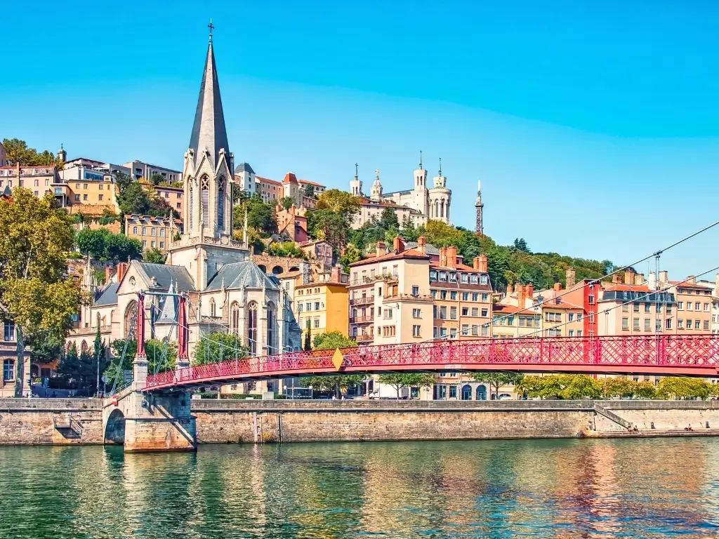 The picturesque Saint-Georges church and red footbridge span the Saône River in Lyon, with the historic hillside district of Fourvière in the background. Lyon, famous for its culinary delights and UNESCO-listed architecture, is a famous day trip from Paris by train.