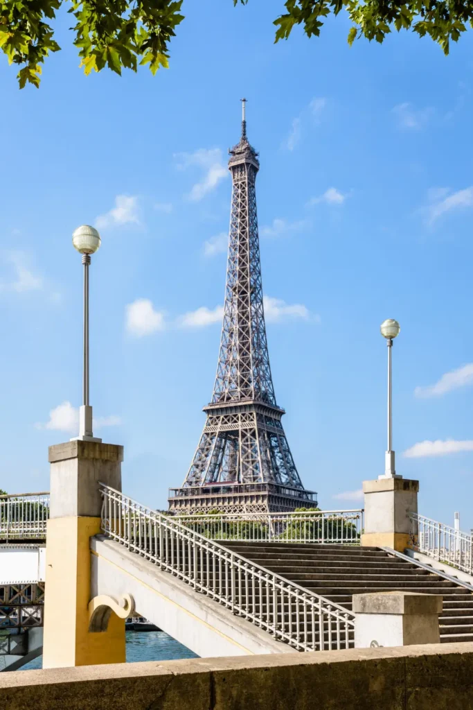Clear view of the Eiffel Tower framed by the elegant arches and lamps of Passerelle Debilly, with lush trees on the banks of the Seine River, on a bright sunny day.