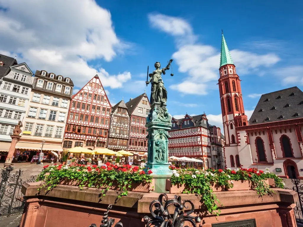 The Römerberg plaza in Frankfurt, Germany, bustling with activity, features traditional half-timbered houses and the Old St. Nicholas Church tower. A statue of Lady Justice foregrounds the scene, with red and white flowers adding a pop of color. Frankfurt is one of the easily accessible day trips from Paris by train
