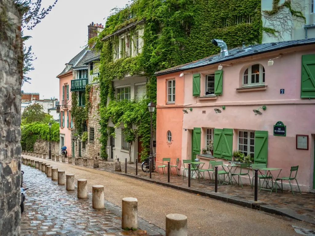 Cobblestone street in Montmartre, Paris, with quaint buildings covered in ivy and a sidewalk café featuring green shutters and matching furniture, conveying a quiet, charming atmosphere.