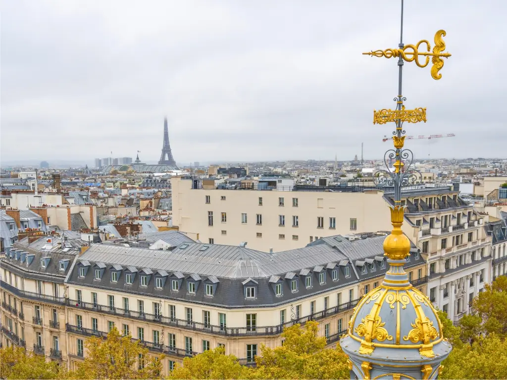 Elevated view of Parisian streets with the gilded dome of the Printemps department store in the foreground and the ornate facade of the Palais Garnier opera house in the distance, set against a backdrop of classic Haussmann-style buildings under a cloudy sky.