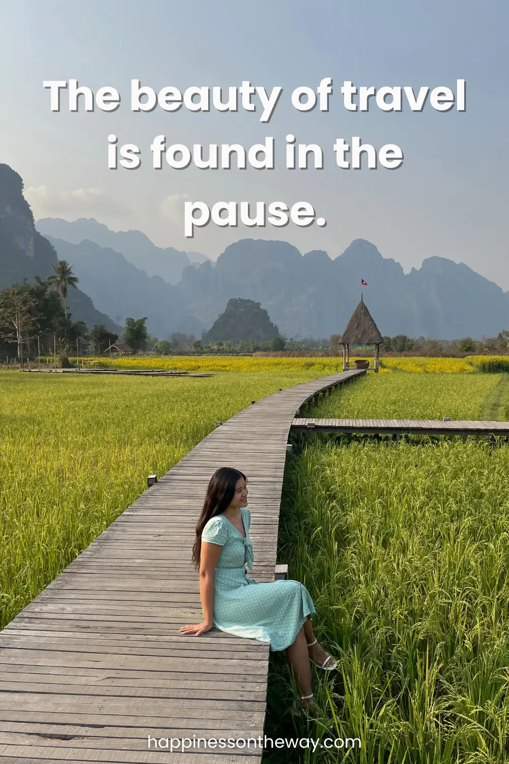 A serene scene capturing slow travel, featuring me in a pastel blue dress sitting on a wooden pathway amidst lush green rice fields with mountains in the distance. The overlaying quote 'The beauty of travel is found in the pause' complements the peaceful landscape, perfect for a slow travel quote.