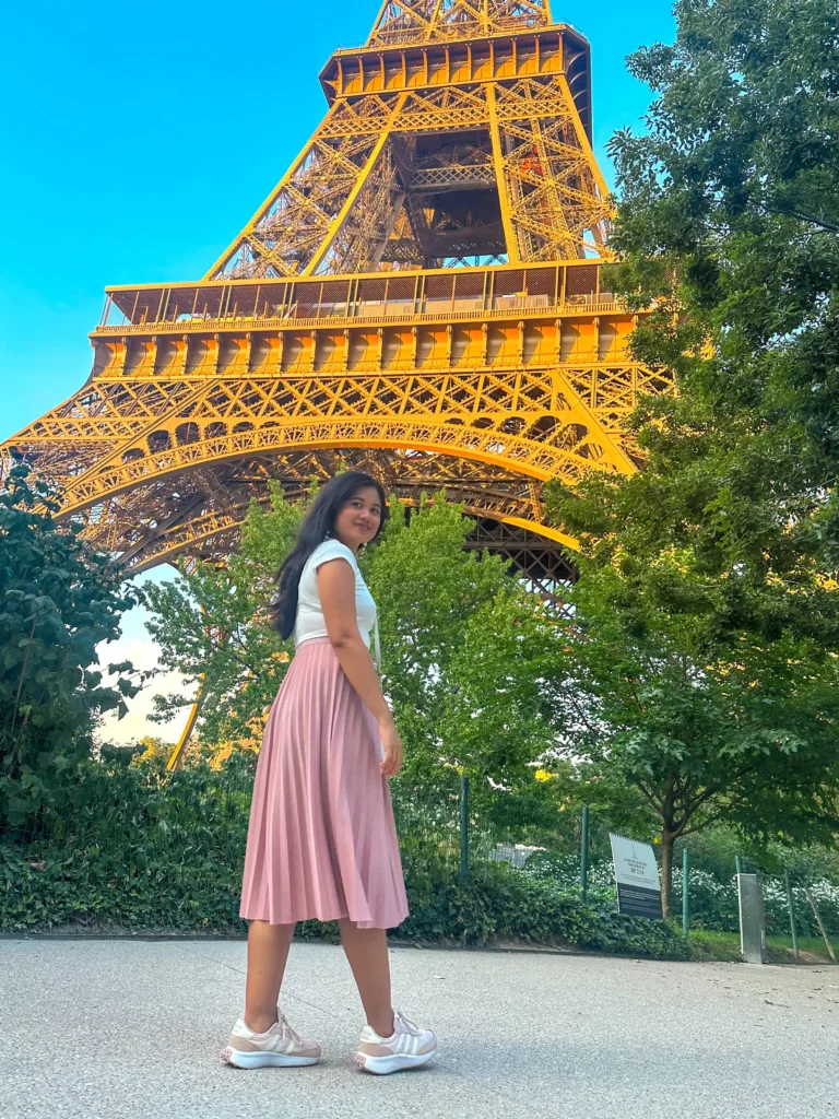Me in white top and pink skirt stands with the Eiffel Tower towering in the background, the lush greenery of the Trocadéro Gardens framing the iconic structure on a clear sunny day.