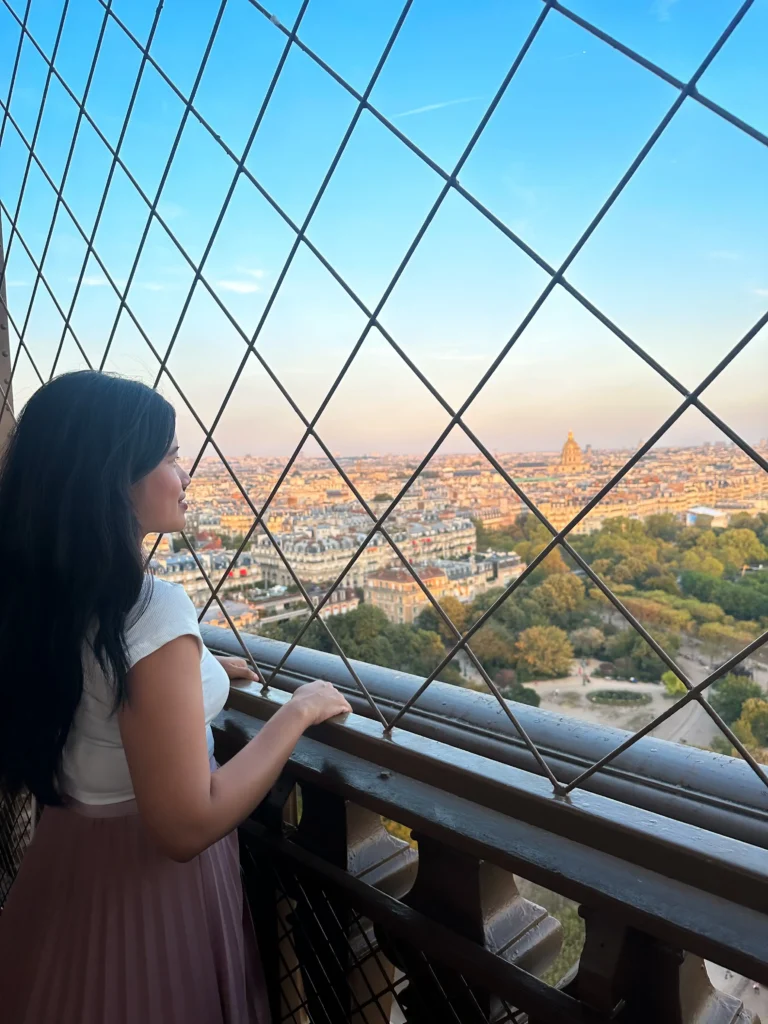 A woman gazes out over the Parisian skyline from the first floor of the Eiffel Tower, with the intricate metalwork of the safety net in the foreground and the city stretching out beneath a sky painted with hues of orange and blue at dusk.