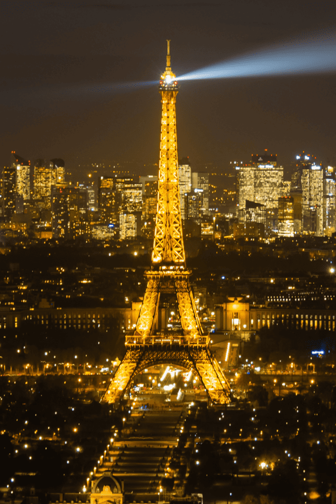 View of Eiffel Tower at night from Tour Montparnasse, with its beacon light piercing the dark sky. The Parisian cityscape glows with lights stretching towards the horizon, highlighting the contrast between the landmark and the urban environment.
