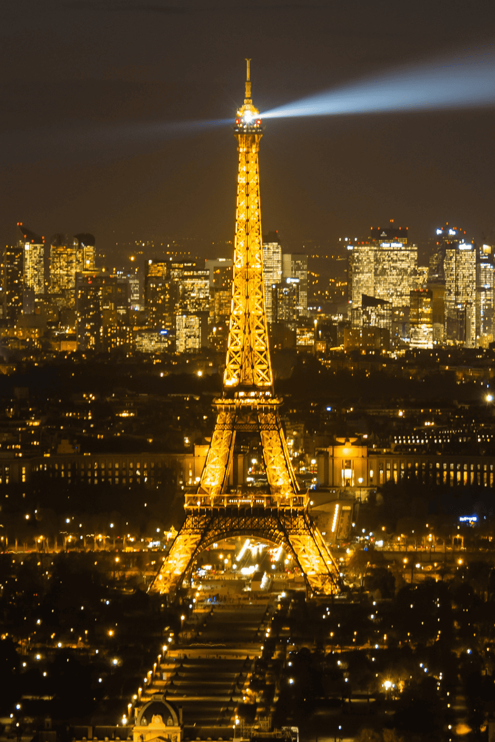 View of Eiffel Tower at night from Tour Montparnasse, with its beacon light piercing the dark sky. The Parisian cityscape glows with lights stretching towards the horizon, highlighting the contrast between the landmark and the urban environment.