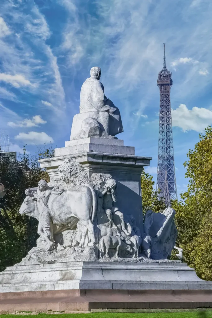 The Eiffel Tower seen from Place de Breteuil, with a striking white statue in the foreground depicting a seated figure and animal figures, set against a vivid blue sky with wispy clouds.