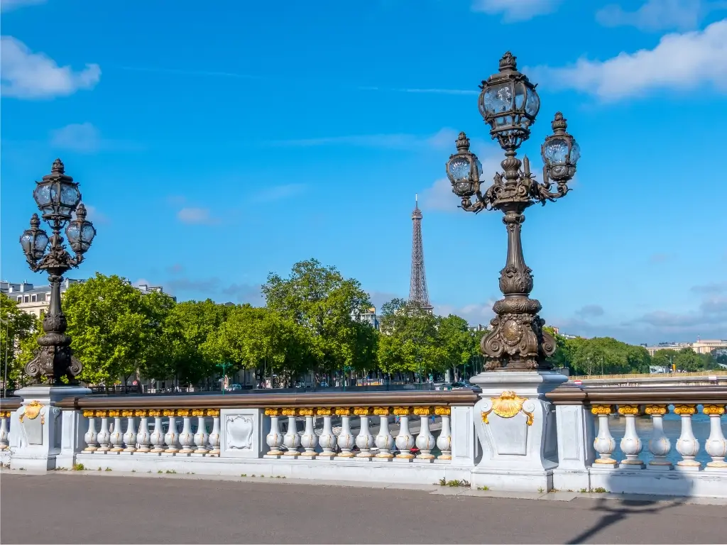 Ornate street lamps on Pont Alexandre III with the view of Eiffel Tower in the background, set against a clear blue sky and lush greenery along the Seine River.