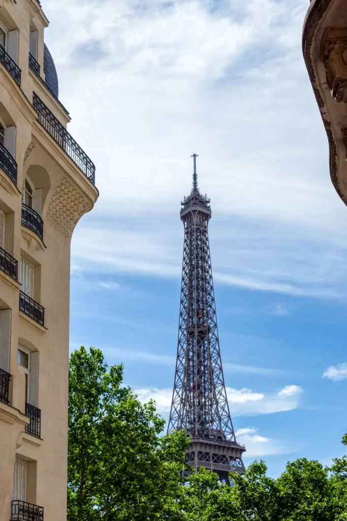 The Eiffel Tower view from Square Rapp, with their balconies and creamy facades contrasting with the lush green treetops and the scattered clouds in the blue sky above.