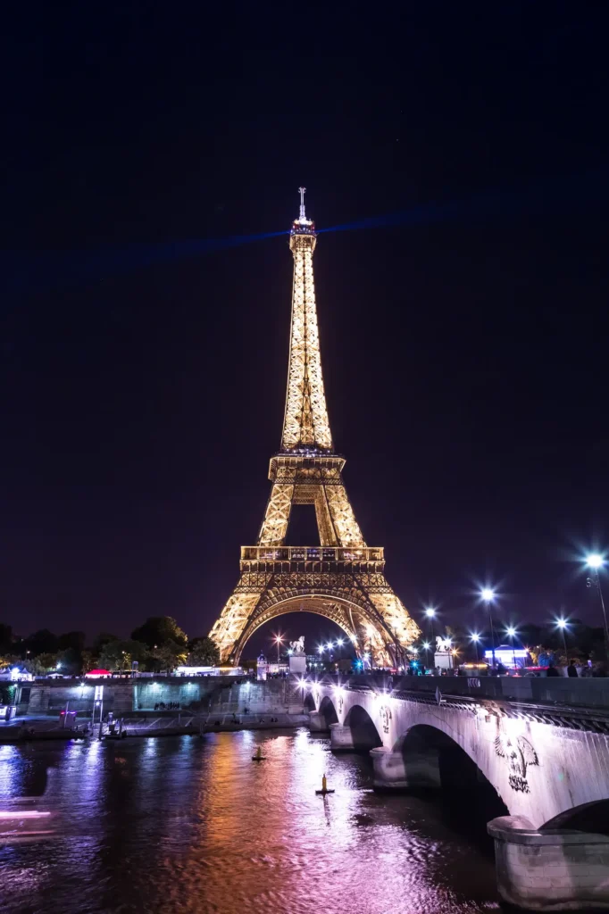 The Eiffel Tower illuminated at night, with its golden lights reflecting on the Seine River, viewed from Pont d'Iéna. The night sky provides a dark backdrop to the vibrant lights and the arching bridge in the foreground adds depth to the urban night scene.