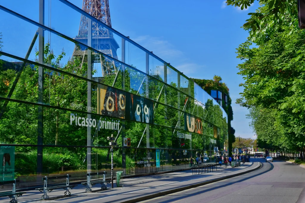 A view from Quai Branly Museum in Paris, showing a vibrant advertisement for a Picasso exhibition on a glass pane with a reflection of the Eiffel Tower, harmoniously blending culture and iconic architecture.