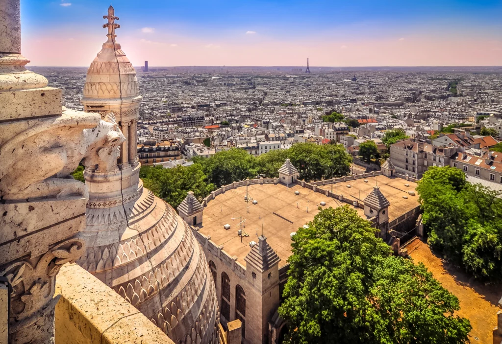 Aerial view from the dome of Sacré-Cœur Basilica in Paris, with a detailed stone gargoyle in the foreground overlooking the cityscape. In the distance, the iconic Eiffel Tower rises above the urban sprawl under a soft blue sky.