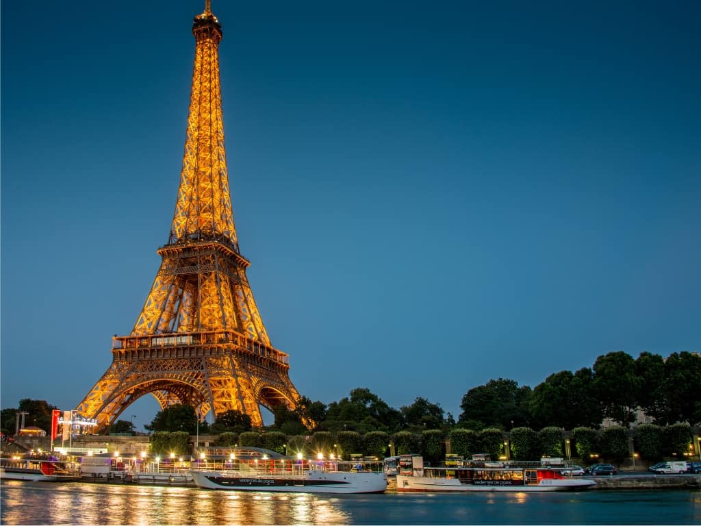 A View of Eiffel Tower illuminated at night, from the Seine River with moored sightseeing boats and tree-lined banks under a twilight sky.