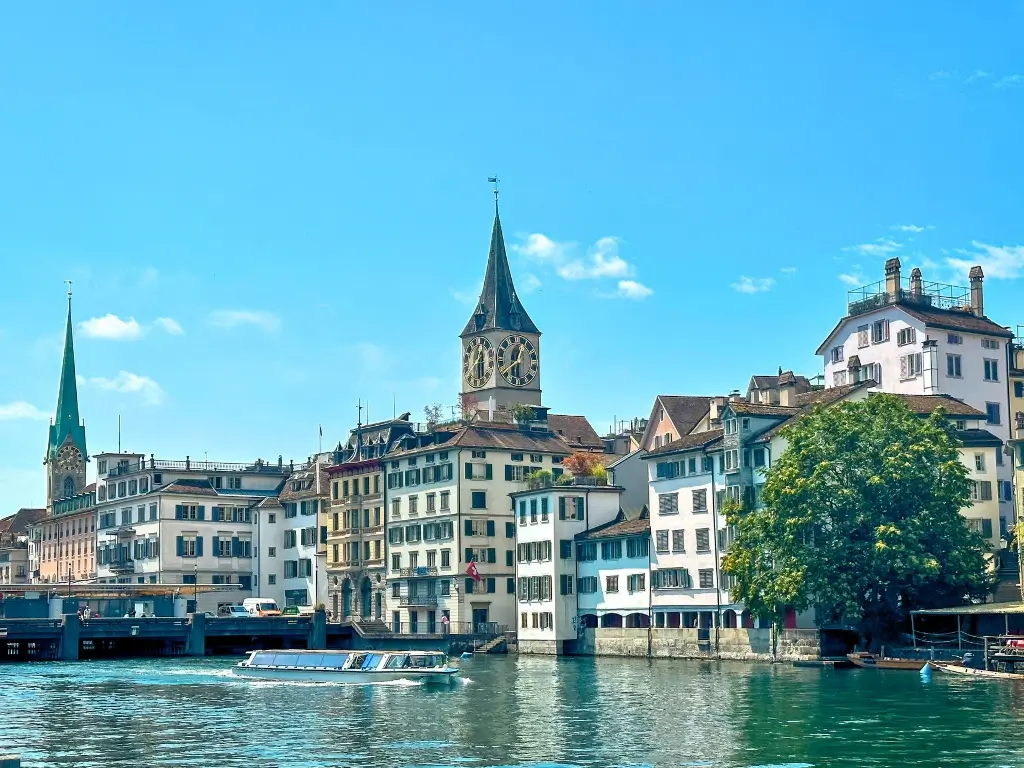 A boat cruising on the Limmat river with the St. Peter church spire in the background, in Zurich, accessible from Paris by train for a day trip.