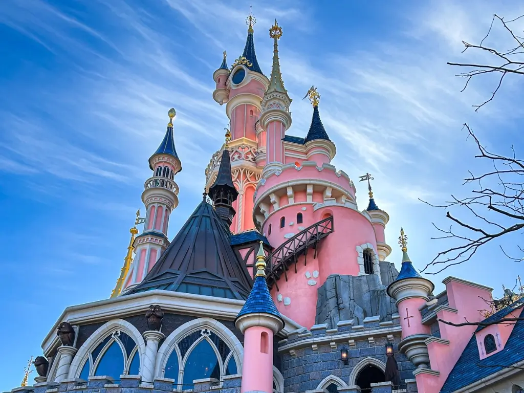 A vibrant photo capturing the iconic Sleeping Beauty Castle at Disneyland Paris, with its pink and blue spires and golden accents, set against a clear blue sky with streaky clouds. Disneyland Paris is among the best day trips from Paris by train.