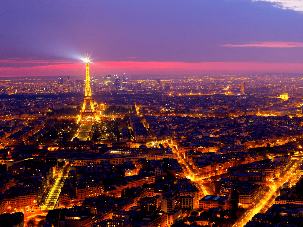 A breathtaking nighttime view of Paris with the Eiffel Tower from Invalides, its beacon shining across the cityscape. The skyline glows under a dusky sky transitioning from sunset to evening, with the city lights tracing the streets below.