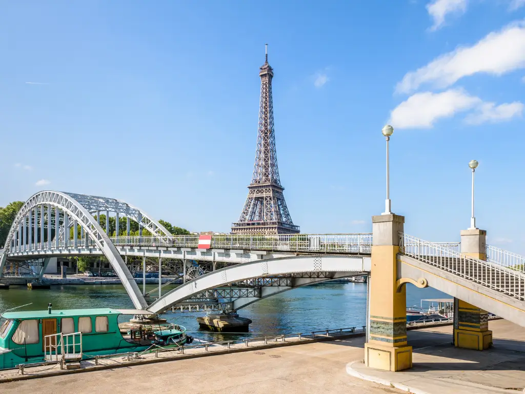 The Eiffel Tower captured from Passerelle Debilly, with the bridge's intricate metalwork and street lamps in the foreground, set against a clear blue sky.