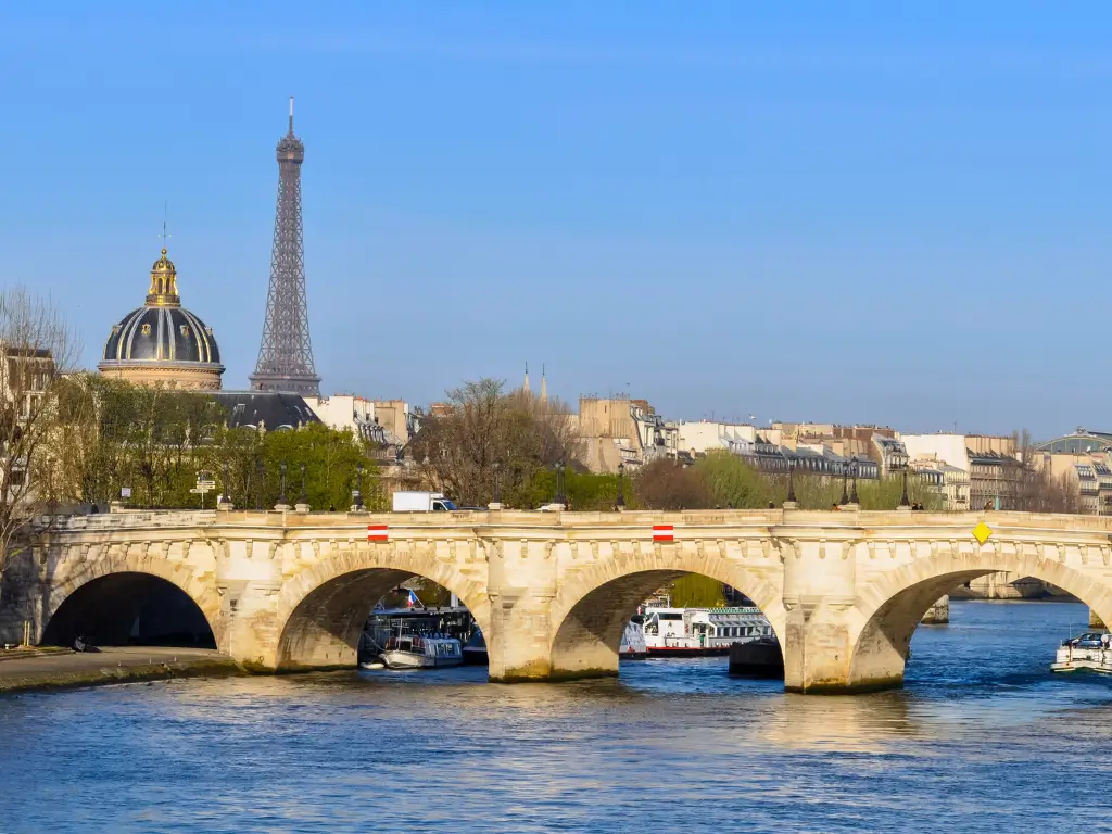The Eiffel Tower and the gold-domed Les Invalides stand tall behind the historic Pont Neuf bridge over the Seine River, with boats gliding on the water, encapsulating a classic Parisian scene.